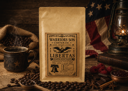A bag of Warriors Son Coffee Co. Libertas coffee stands upright on a rustic wooden surface, surrounded by coffee beans, a mug, a lantern, and an American flag in the background.