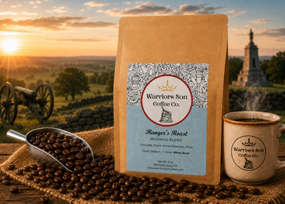 A bag of Warrior Son Coffee Co. Ranger's Roast medium roast coffee stands on a wooden surface with coffee beans spilled around it, alongside a ceramic mug and a metal scoop of beans.
